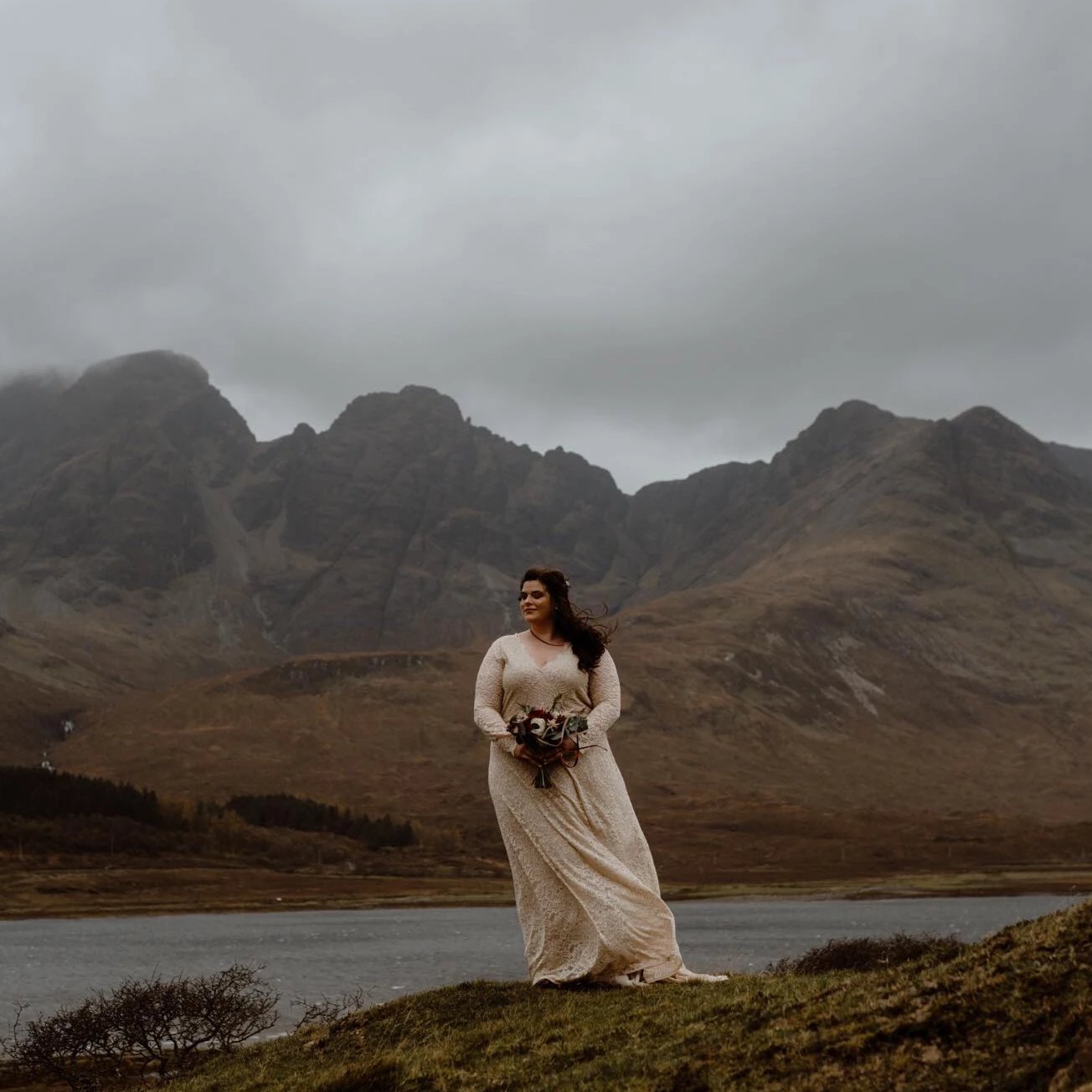 Bride in a champagne lace wrap wedding gown with long sleeves standing beside a mountain lake.