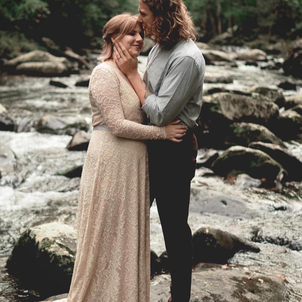 Bride in a champagne lace wrap wedding gown with long sleeves embracing groom beside a rocky stream. 