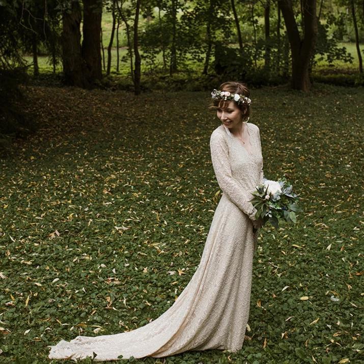 Bride in a champagne lace wrap wedding gown with long sleeves holding flowers in a forest setting.