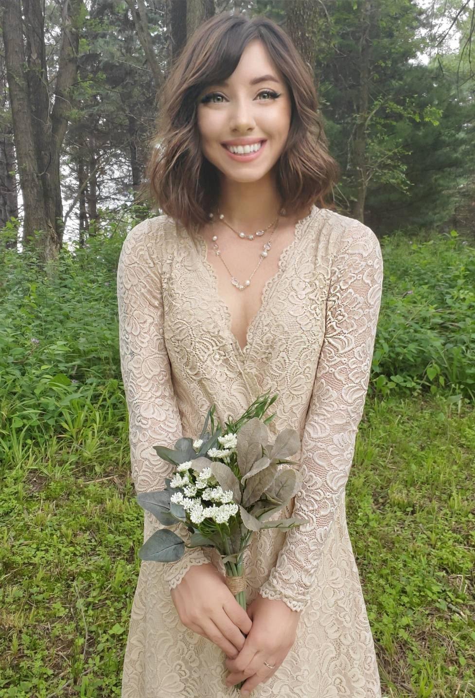Bride in a champagne lace wrap wedding gown with long sleeves holding flowers in a forest setting.