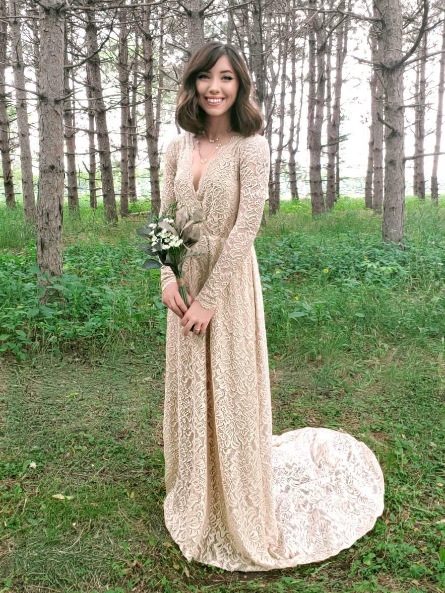 Bride in a champagne lace wrap wedding gown with long sleeves standing in a forest setting.