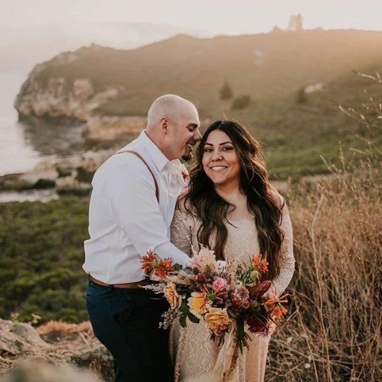 Bride and groom in a romantic side on cuddle in an outdoor coastal wedding portrait, bride holding a bouquet and wearing a champagne long-sleeve lace wedding gown.