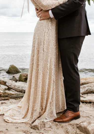 Close-up detail of bride wearing a champagne lace wedding gown beside the groom in a coastal bridal setting, showing the flowing skirt and train.