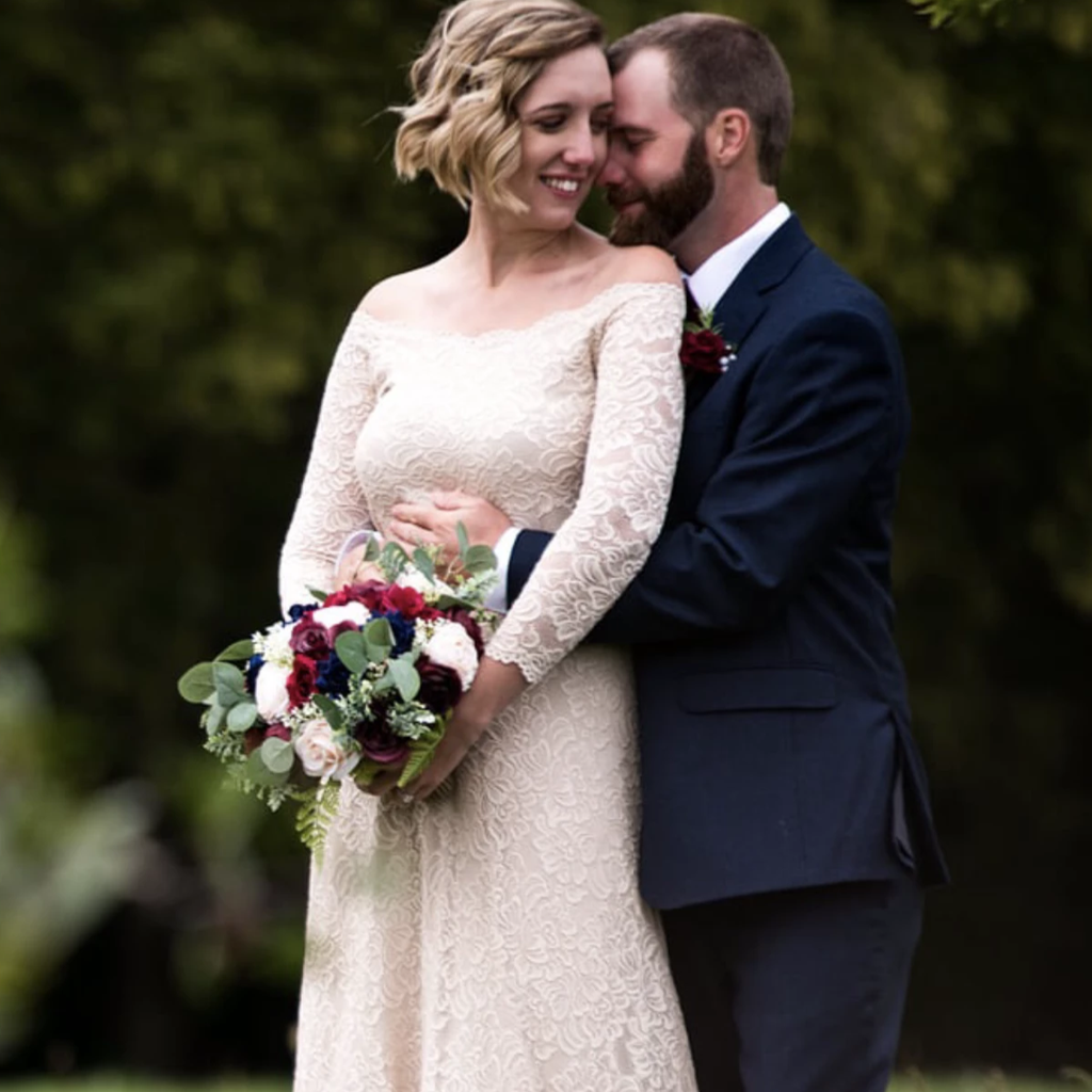 Bride holding a bouquet being held from the rear by the groom in a romantic outdoor wedding portrait, wearing a champagne lace wedding gown with long sleeves.