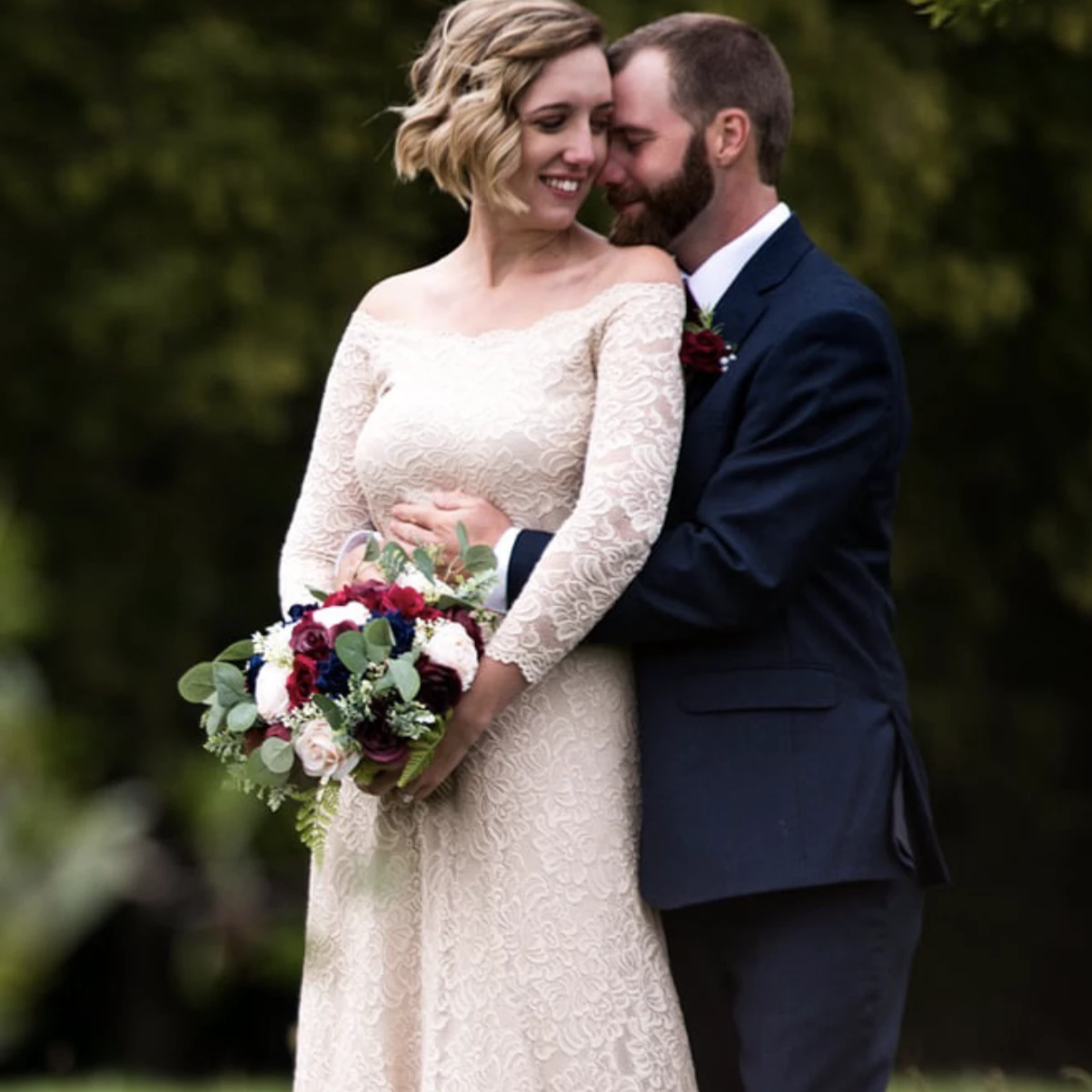 Bride holding a bouquet being held from the rear by the groom in a romantic outdoor wedding portrait, wearing a champagne lace wedding gown with long sleeves.