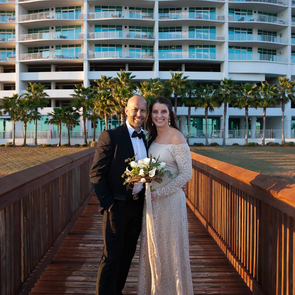 Bride and groom posing on a wooden resort walkway in a destination wedding setting, bride wearing a champagne long-sleeve lace wedding gown and holding a bouquet.