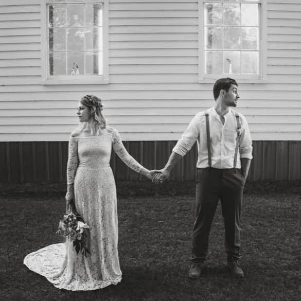Bride and groom holding hands beside a rustic building, bride wearing a champagne off-shoulder lace wedding gown with long sleeves, fitted silhouette, and train.