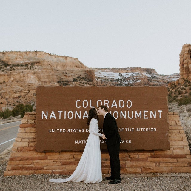 Bride and Groom kissing in front of the Colorado National Monument sign, bride wearing an Ivory lace wedding gown with a long sleeve gown and a flowing train.