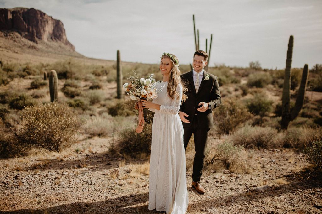 Bride with bouquet of flowers and groom posing for photo's together in an Arizona desert landscape with saguaro cacti. The bride wearing a Ivory open back lace wedding gown with high collar neckline, long lace sleeves and flowing floor-length skirt "