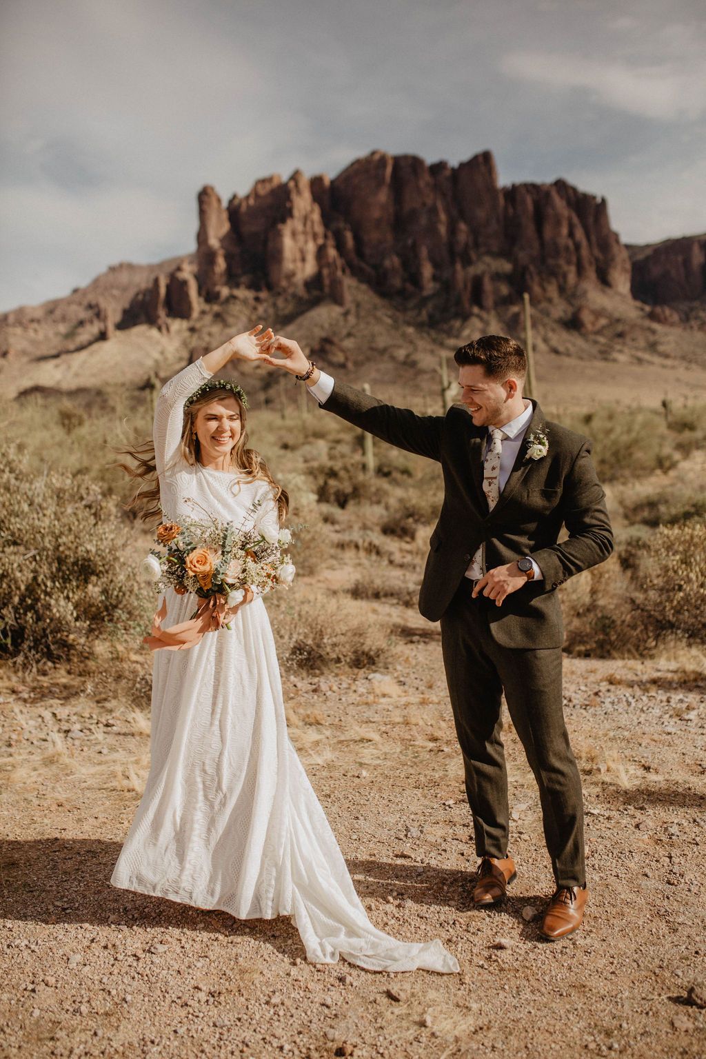 Front view of Bride with peach bouquet and groom dancing joyfully in a desert landscape with canyon backdrop — bride wearing an Ivory open back lace wedding dress with high collar neckline, flowing full-length skirt and flowing and train.