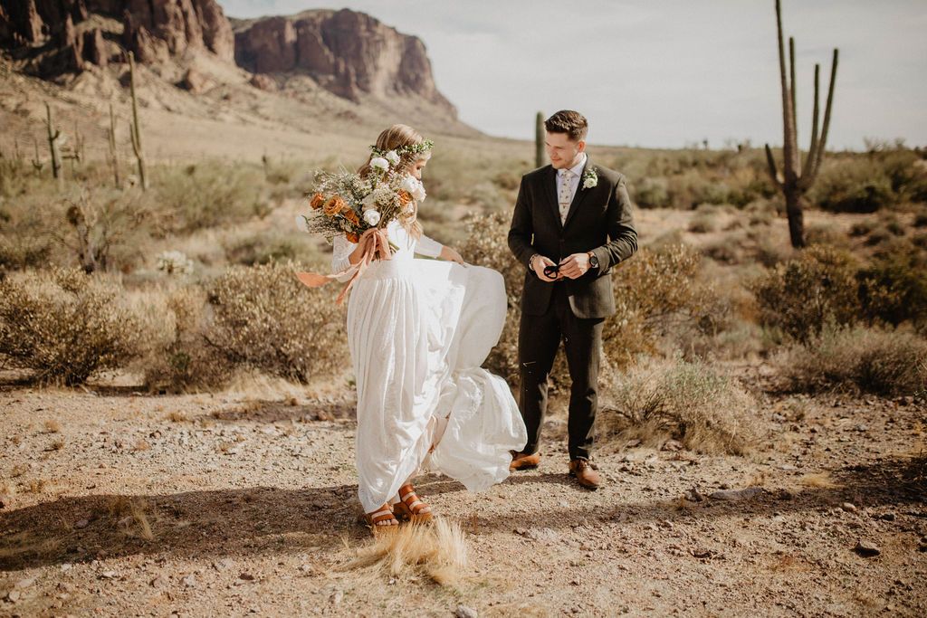 Bride playfully lifting the flowing skirt of her lvory long-sleeve lace wedding gown beside the groom in a desert wedding setting with a sagaro cacti.