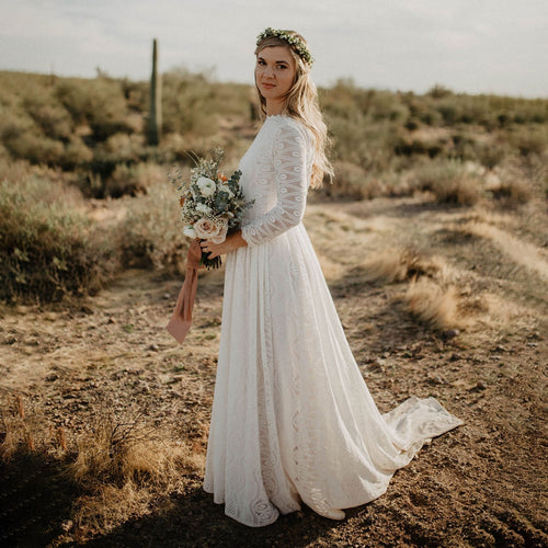 Left side view of bride standing in a desert landscape wearing an Ivory open-back lace wedding gown with high collar neckline, long geometric lace sleeves, A-line floor-length skirt  flowing and with  train visible —Curated by Douceur de Vivre Paris.