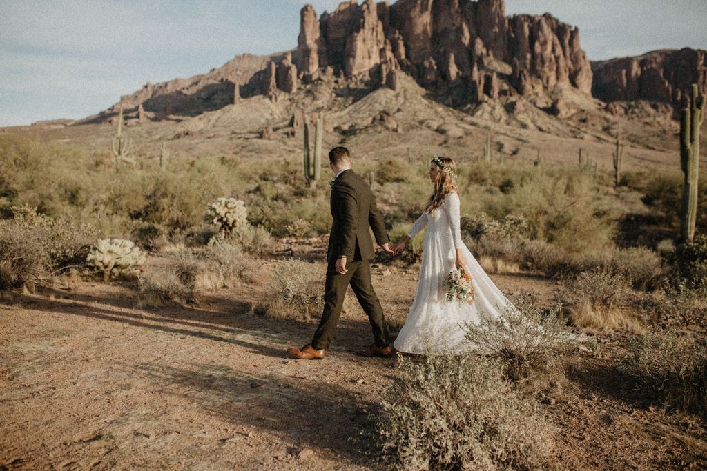 Bride and groom walking hand in hand through a desert canyon wedding setting, bride wearing an ivory long-sleeve lace bridal gown with open-back detail and flowing train.