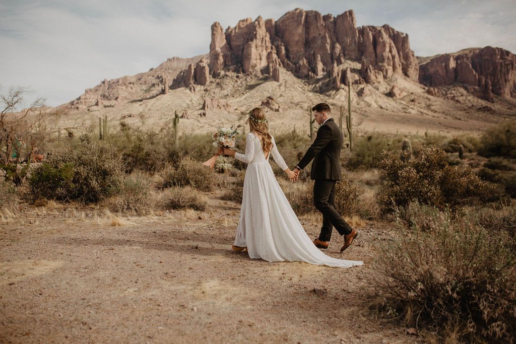 Bride and groom walking together in a desert canyon wedding setting, with the bride wearing an ivory long-sleeve lace bridal gown and flowing train.