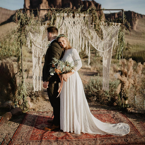 Left sideview of bride and groom embracing under a macramé arch in a desert canyon setting — bride wearing a white open back lace wedding dress with long sleeves, floor-length skirt and train — Curated by  Douceur de Vivre Paris.