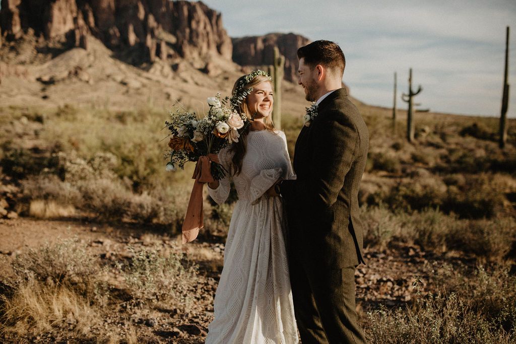 Bride holding a bouquet and smiling at the groom in a desert setting, wearing an Ivory long-sleeve lace bridal gown with a collar neckline and a flowing dress.