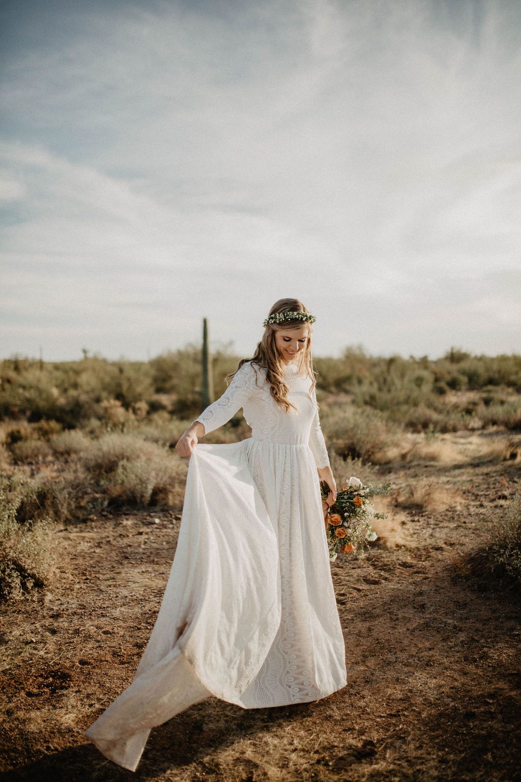 Frontview of bride walking outdoors in a desert setting lifting her ivory open back lace wedding gown with a collar neckline, long lace sleeves, flowing floor-length skirt, and train.