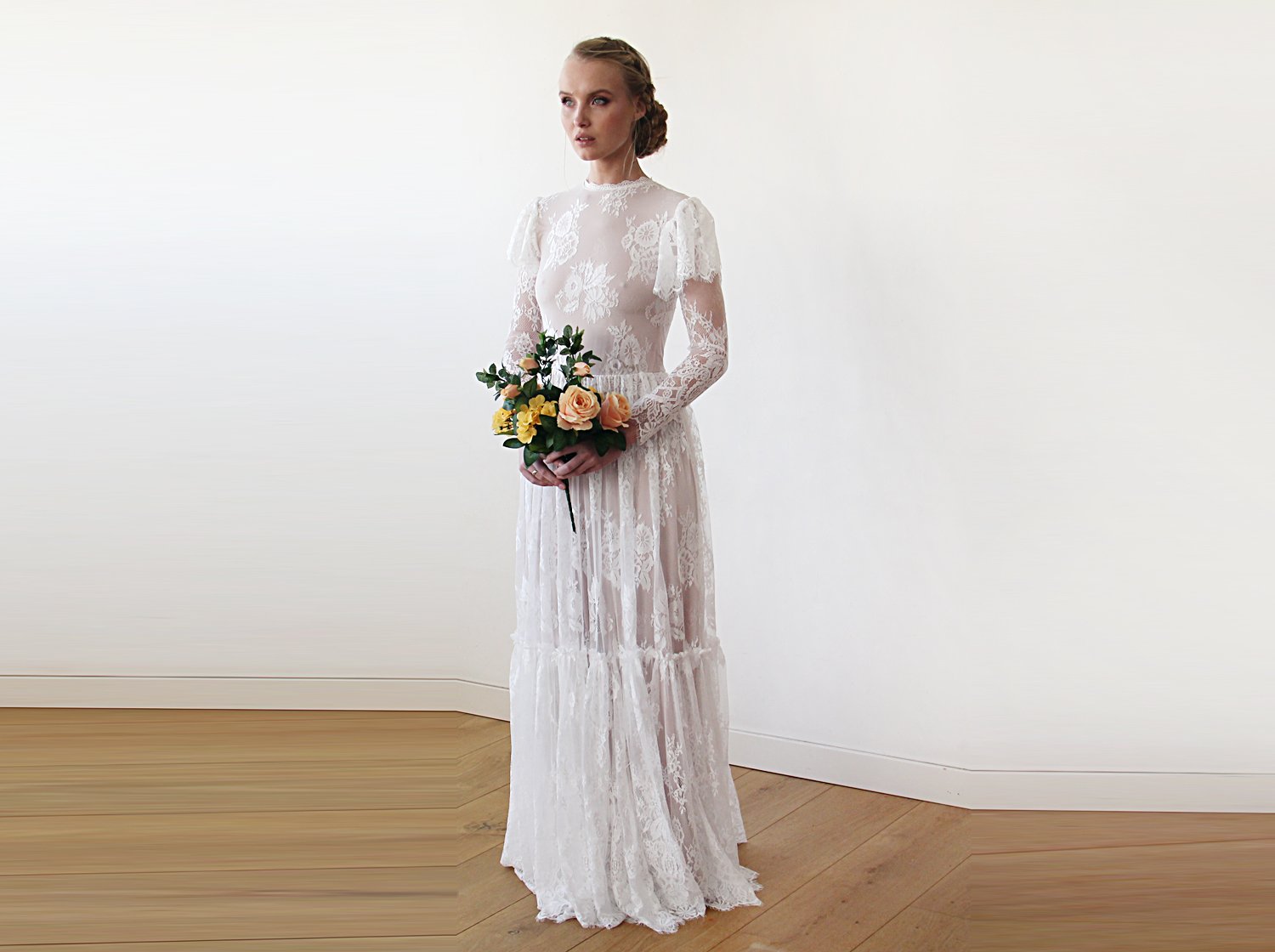 Full-length front view of model with a bridal bouquet wearing an ivory woodland vintage lace wedding gown with high neckline, sheer long lace sleeves, flutter sleeve overlay, tiered skirt, and bridal bouquet.