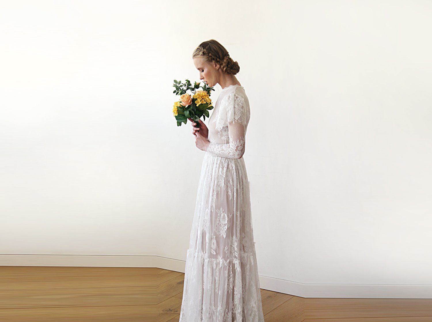 Left side full-length side view of model with a bridal bouquet wearing an ivory woodland vintage lace wedding gown with high neckline, flutter sleeve overlay, sheer long lace sleeves, tiered skirt, and bridal bouquet.